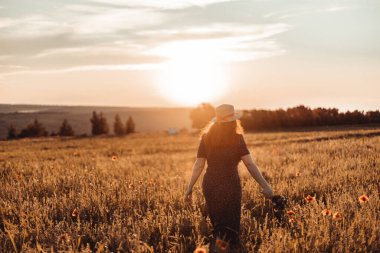 Bir buket kır çiçeğiyle yaz elbisesi içinde mutlu bir kadın tarlada sıcacık bir günbatımının tadını çıkarıyor.