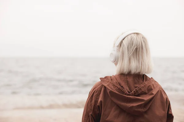 Widok z tyłu blondynki noszącej słuchawki słuchające muzyki lub podcast z aplikacji smartfona przeciwko morzu — Zdjęcie stockowe