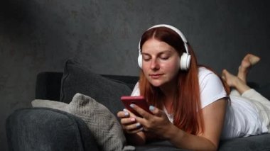 Rahatlamış genç bir kadın evde kanepede yatarken akıllı telefonuyla müzik dinliyor..