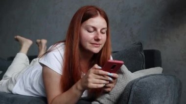 30 'lu yaşlarda, güzel, kızıl saçlı, kanepede yatan ve evinde akıllı telefon kullanan bir kız. Sosyal medyayı izliyordum. Teknoloji iletişim konsepti