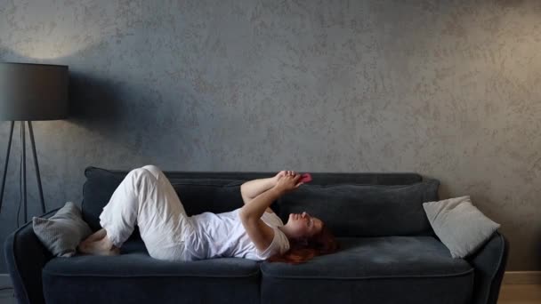 Relaxado bela jovem usa um smartphone desfrutando fim de semana no sofá acolhedor em casa sala de estar — Vídeo de Stock
