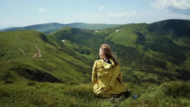Gyönyörű szőke nő turista világos sárga kabátban ül a hegy tetején, és nézi Mountain Valley. Arany haj a friss levegőben szél — Stock videók
