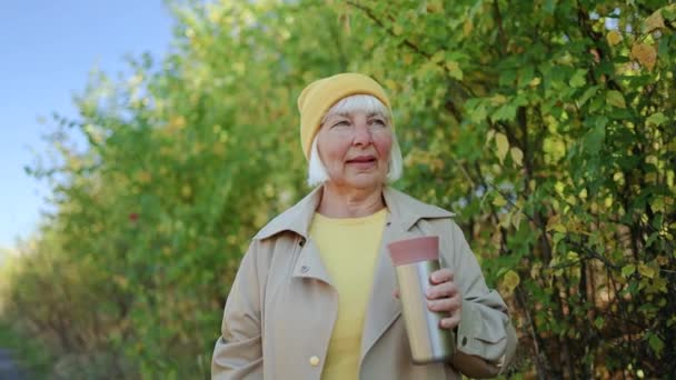Schöne blondhaarige Senioren in stylischer Kleidung trinken einen heißen Kaffee, genießen die Natur und gehen im herbstlichen Park spazieren — Stockvideo