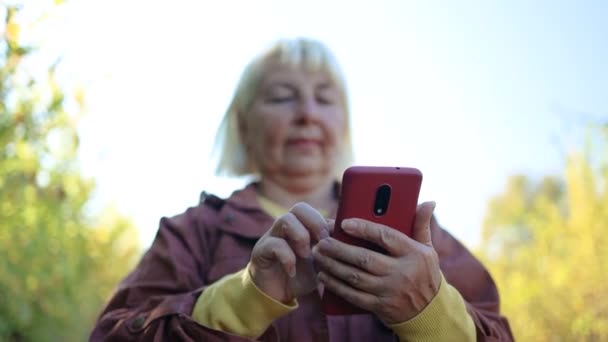 Portrét šťastné krásné blond zralá žena ve stylovém oblečení pomocí mobilního telefonu se těší přírodě v podzimním městském parku. 5G technologické zařízení. — Stock video