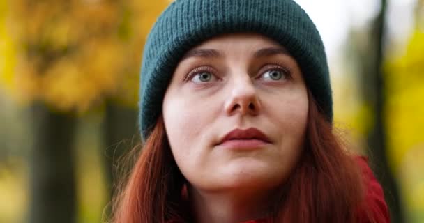 Egy gyönyörű fiatal lány, 30 éves, zöld meleg kalapban és piros kabátban, az őszi arany erdő kamerájába néz. — Stock videók