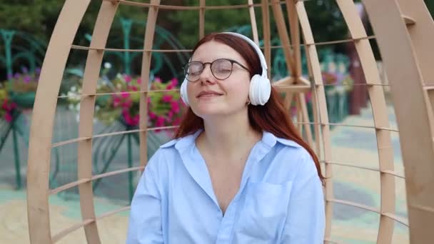 Caucasienne belle jeune femme prenant des écouteurs et écoutant de la musique à l'extérieur au parc de la ville au coucher du soleil — Video