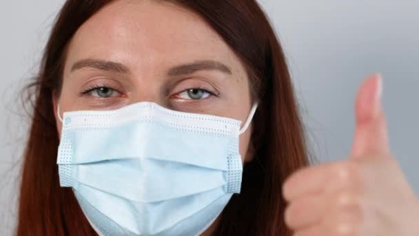 COVID-19 로부터 보호하기 위해 마스크를 쓰고 회색 배경에서 고립된 엄지손가락을 내밀고 있는 젊은 여성 — 비디오