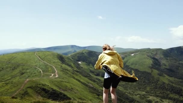 Жінка - блондинка, що подорожує в яскраво-жовтій куртці на вершині гори і дивиться на гірську долину в літній сонячний день. Золоте волосся у свіжому повітрі. — стокове відео