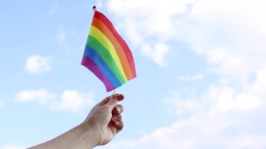 Dişi el açık mavi bir arka plana karşı küçük renkli bir gökkuşağı gay gurur bayrağı sallıyor.