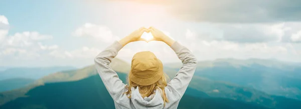 Vista posterior de la joven viajera hembra en sombrero de pie sobre los picos de montaña de verano y haciendo forma de corazón de amor por las manos. —  Fotos de Stock