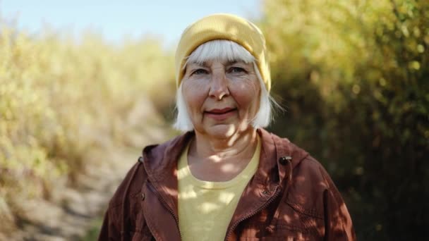 Retrato de una hermosa mujer mayor rubia feliz con ropa elegante sonriendo a la cámara caminando sola en el parque durante el otoño — Vídeo de stock