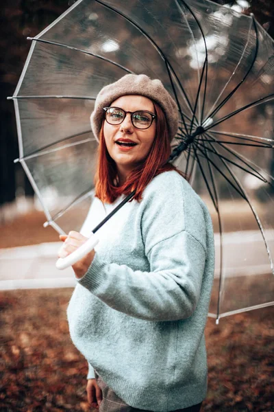 Όμορφη κοκκινομάλλα κοπέλα με γυαλιά και βιβλία για μελέτη κοίτα την κάμερα στο πάρκο. Διαφανής ομπρέλα από τον άνεμο και τη βροχή σε κακές καιρικές συνθήκες — Φωτογραφία Αρχείου