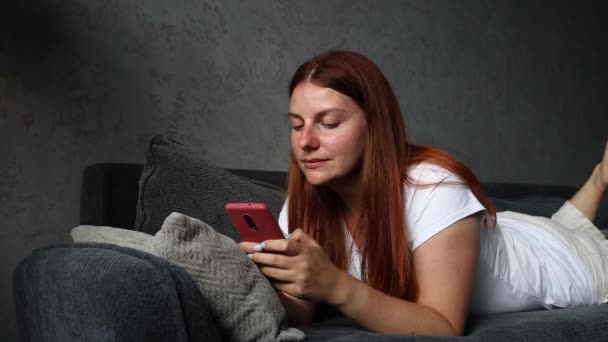 Fille souriante aux cheveux roux couchée sur le canapé et utilise un smartphone. Les travailleurs indépendants utilisent les téléphones mobiles pour lire les informations — Video