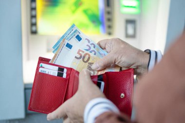 Tanınmayan bir kadın, caddedeki ATM 'nin yanındaki kırmızı deri bir cüzdandan 50 ve 20 avroluk banknot çıkarıyor..