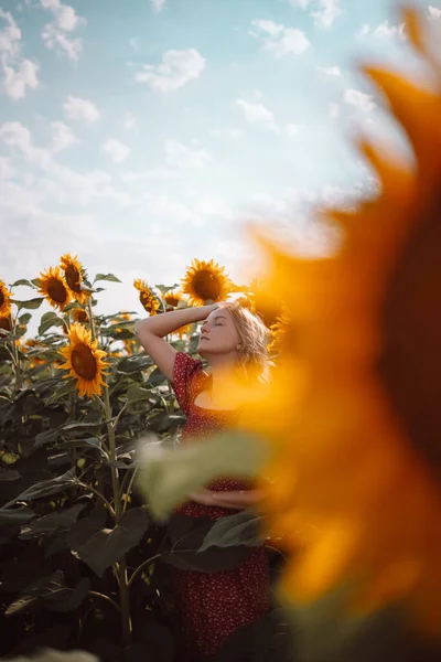 Adorabile ragazza caucasica posa in un campo di girasole alla luce del sole. Donna in abito rosso stile estivo con le mani aperte sopra cielo blu nuvoloso — Foto Stock