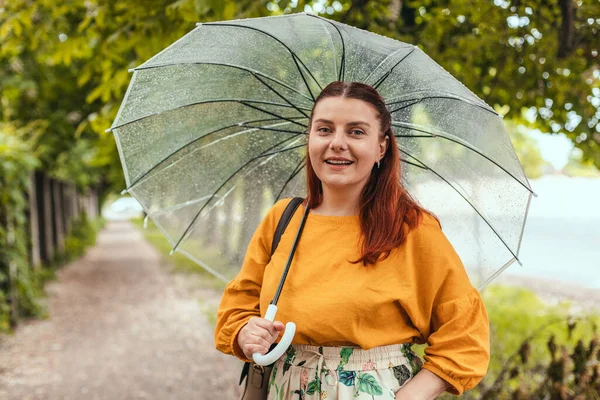 Κομψή νεαρή γυναίκα με φωτεινά ρούχα κάτω από μια διάφανη ομπρέλα σε κακές καιρικές συνθήκες στο πάρκο της πόλης — Φωτογραφία Αρχείου