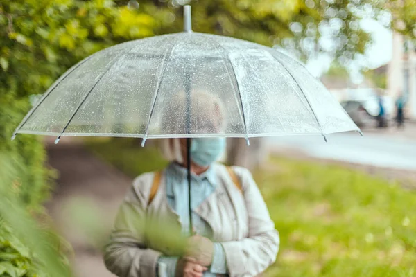 Felnőtt nő fehér kalapban és meleg sállal egy átlátszó esernyő alatt egy őszi parkban a szabadban, hideg esős napokon — Stock Fotó