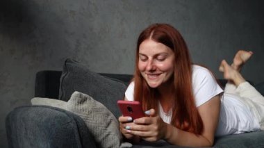 Güzel kızıl saçlı, gülümseyen kız kanepede uzanıyor ve evinde akıllı telefon kullanıyor. Sosyal medyayı izliyordum. Teknoloji iletişim konsepti