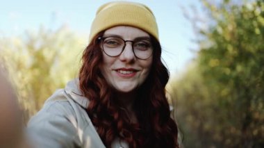 Güzel kıvırcık saçlı şık elbiseli ve gözlüklü kız kameraya gülümseyip gülüyor, doğanın tadını çıkarıyor ve sonbahar parkında yürüyor.