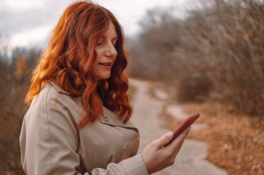 Genç bir kadın internette sohbet etmek ve şehir parkında yürümek için akıllı telefon kullanıyor.