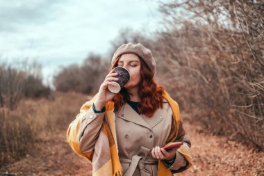 Akıllı telefon kullanan ve şehir parkında kahve içen güzel genç bir kadın.