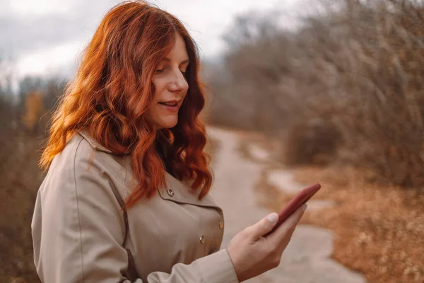 Genç bir kadın internette sohbet etmek ve şehir parkında yürümek için akıllı telefon kullanıyor.