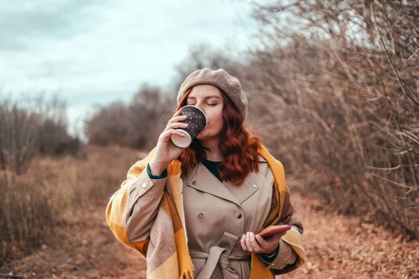 Akıllı telefon kullanan ve şehir parkında kahve içen güzel genç bir kadın.
