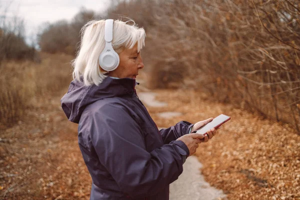 在城市的秋天公园里 中年美丽的金发女子正在用耳机听音乐 — 图库照片