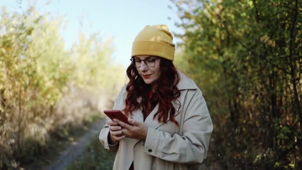 Hermosa mujer rizada de pelo rojo con ropa y gafas brillantes y elegantes utiliza un teléfono inteligente, escribe SMS y disfruta de la naturaleza en un parque de otoño de la ciudad. Concepto de tecnología y felicidad. — Vídeo de stock