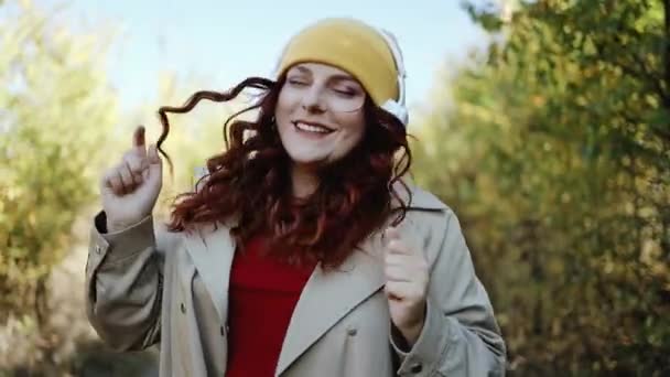 Joven mujer hipster feliz divirtiéndose, escuchando música y bailando en la calle al aire libre, parque de otoño de la ciudad. Mujer con el pelo rizado en ropa elegante disfrutando de la música — Vídeo de stock