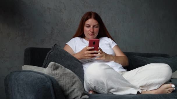美しい若い30代の赤い髪の笑顔の女の子がソファの上に横たわっており、自宅のリビングルームでスマートフォンを使用しています。ソーシャルメディアを見ている。技術コミュニケーションの概念 — ストック動画