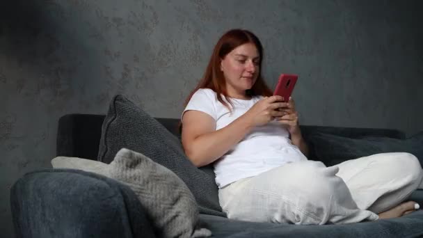 Les mains féminines tiennent le téléphone intelligent couché sur le canapé et utilisent un téléphone dans le salon de la maison. Regarder les médias sociaux. Concept de communication technologique — Video