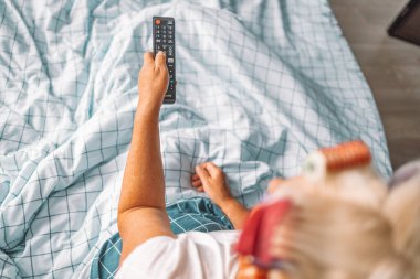 Kadın beyaz bir battaniyenin altında yatakta otururken televizyon kanallarını değiştiriyor. Yukarıdan görüntüle