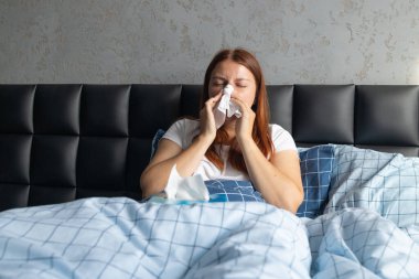 Mendili akan, burnu akan, soğuk enfeksiyonu olan, depresyonda ve stresli bir kadın yatak odasında oturuyor.