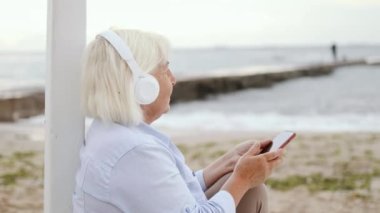 Sarışın kadın kulaklık takıyor, meditasyon yapıyor, müzik dinliyor ya da denize karşı akıllı telefon uygulamasından podcast yapıyor.