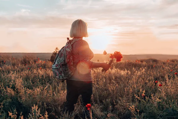 Вид сзади женщины туристки с рюкзаком и маковым цветочным букетом на закате в поле. Летние каникулы. Счастливая девушка наслаждается солнцем. Горизонтальное знамя — стоковое фото