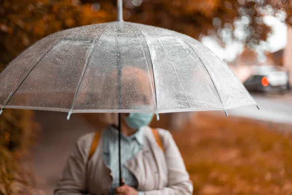 Felnőtt nő fehér kalapban és meleg sállal egy átlátszó esernyő alatt egy őszi parkban a szabadban, hideg esős napokon — Stock Fotó