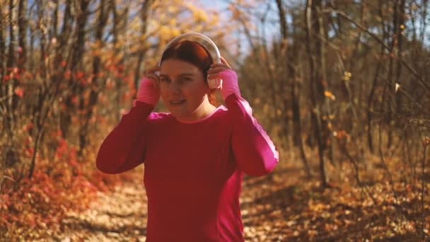 Активная девушка в спортивной одежде надевает беспроводные наушники, чтобы начать бег трусцой по улице в парке осеннего леса на природе — стоковое видео