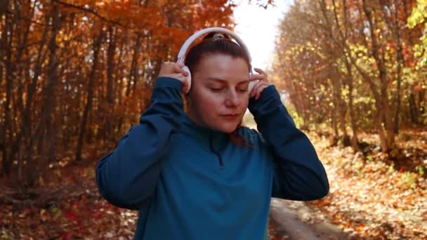 在大自然秋天的森林公园里，身穿运动服、头戴白色无线耳机的活跃的女运动员在户外跑步。锻炼、福利和体育女童的概念 — 图库视频影像