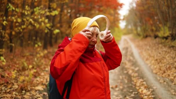 Active Caucasian 40s 50s female walking along, having fun with headphones mobile phone and backpack enjoying listening to music outdoors in autumn city park — Stock Video