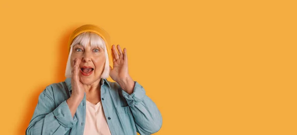 Alegre mujer de los años 50 pelo rubio posando aislado sobre fondo amarillo anaranjado. Gritando y sosteniendo sus manos cerca de su cara —  Fotos de Stock