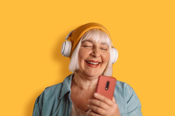 Happy 50s žena sluchátka poslouchat hudbu zpívat píseň záznam hlas od mobilního telefonu diktafon na žluté barvy pozadí studio portrét. — Stock fotografie