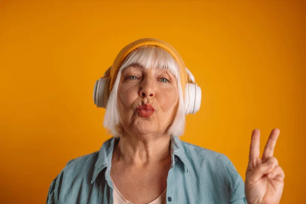 Happy 50s stylish woman giving an air kiss with headphones listening to music and showing peace gesture with hand, enjoying cool playlist — Fotografia de Stock