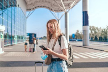 Sırt çantası ve bavulu olan turist kız uluslararası havaalanı yakınında cep telefonu kullanıyor.