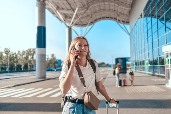 Sırt çantası ve bavuluyla gezgin bir kadın uluslararası havaalanı yakınında cep telefonuyla konuşuyor.