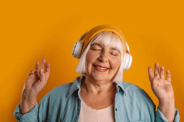 Carefree blonde hair 50s woman with closed eyes dancing, listening to a song in wireless headphones — Fotografia de Stock