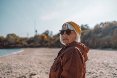 Mutlu, orta yaşlı, güneş gözlüklü kadın deniz kenarında yürürken suya bakmayı seviyor.