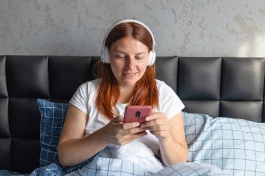 Mutlu kadın yatakta cep telefonu kullanıyor. Kulaklık takan ve akıllı telefondan müzik dinleyen bir kadın.