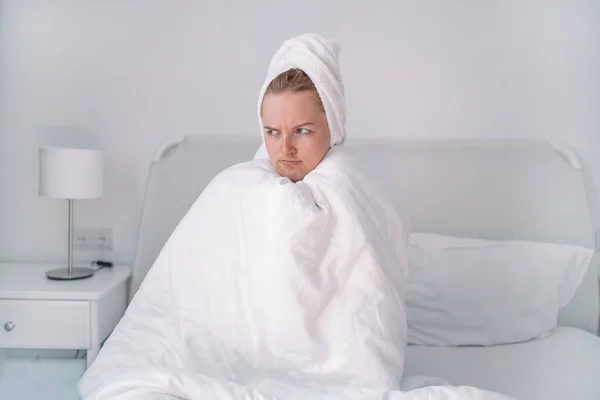 Displeased young blonde girl sitting wrapped in a warm blanket on the bed with an unhappy face, feel sick while sit on bed in bedroom. — Stock Photo, Image