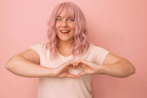 魅力的な若い白人ピンクの髪の女性はピンクの背景に対して幸せとロマンチックな形のハートジェスチャーポーズを感じます — ストック写真
